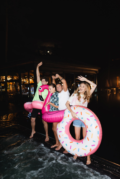 Amigos bailando con anillos de natación durante la fiesta cerca de la piscina por la noche  - Foto, Imagen