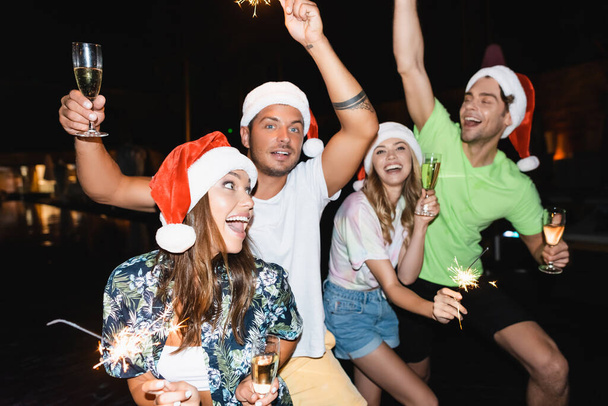 Selective focus of friends in santa hats holding glasses of champagne and sparklers at night  - 写真・画像