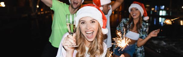 Horizontal image of excited woman holding glass of champagne and sparkler while celebrating new year at night outdoors  - Foto, Imagen