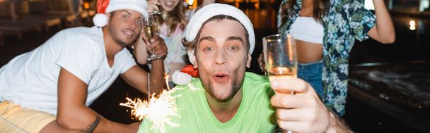 Colpo panoramico di uomo in cappello rosso di Babbo Natale con sparkler e bicchiere di champagne vicino agli amici di notte  - Foto, immagini