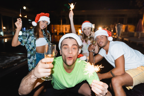 Selective focus of man in santa hat holding sparkler and glass of champagne near friends during party at night  - Photo, Image