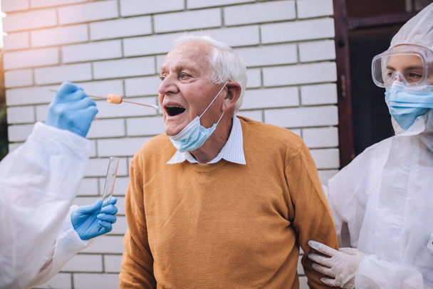 Koruyucu kıyafetli doktorlar, yaşlı bir adamdan koronavirüs enfeksiyonu için örnek alıyorlar. Ev konseptinde Corona virüsü testi örneği alıyoruz, karantinadaki son sınıf öğrencisi.. - Fotoğraf, Görsel