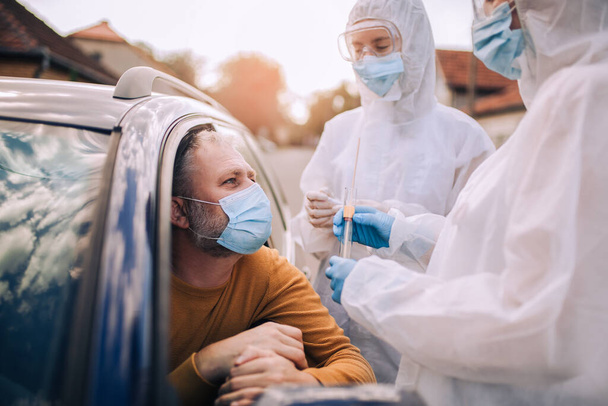 Suojapukuiset lääkärit ottavat näytteen henkilöltä mahdollisen koronavirusinfektion testaamiseksi - Valokuva, kuva
