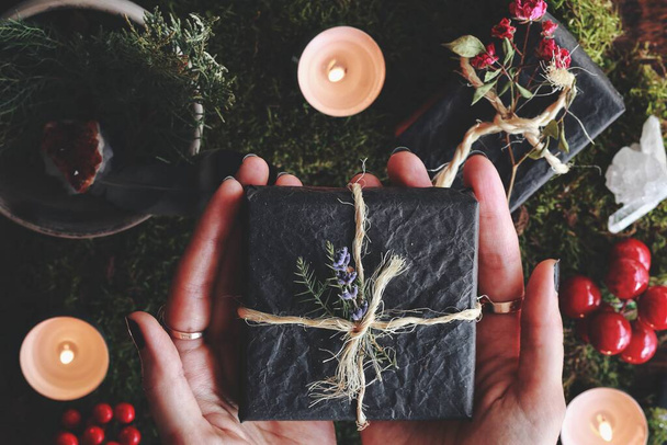 Yule winter solstice (Christmas) themed flat lay of female hands holding a black gift box wrapped in yarn and flowers in her hands. Forest moss, evergreens, burning lit candles, presents, red berries - Foto, imagen