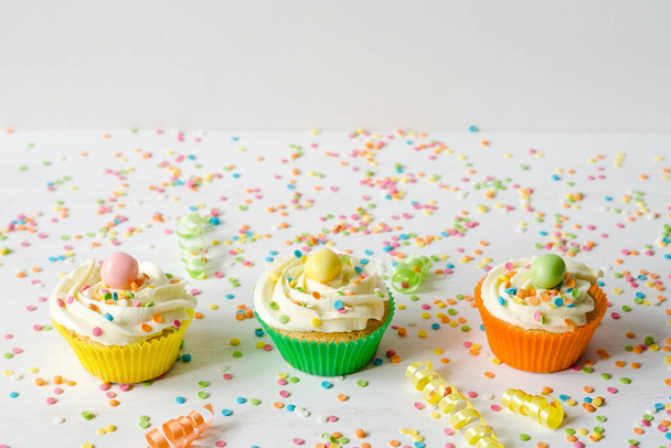 Three brightly colored homemade cupcakes on a white wooden background with sprinkles. Festive birthday concept. Copy space composition - Foto, afbeelding