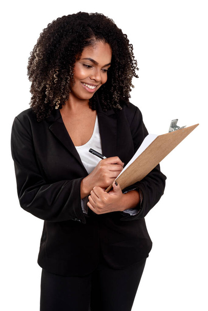 Neşeli ve gülümseyen yönetici siyah Brezilyalı kadın beyaz arka planda izole edilmiş, elinde bir kalem ve bir not panosuyla çalışıyor.. - Fotoğraf, Görsel