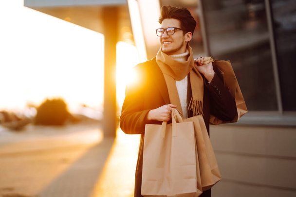 Stylish man with paper packages after shopping at sunset. Handsome man doing shopping in the city. Consumerism, shopping, sales, lifestyle concept. Black friday. - Foto, afbeelding
