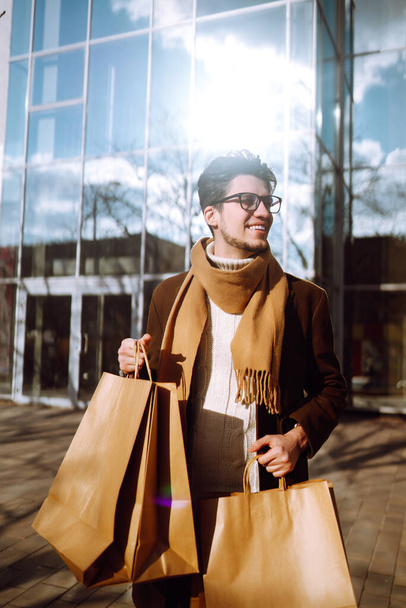 Stylish man with paper packages after shopping. Young man with shopping bags walking around the city after shopping. Black friday, sale, consumerist, lifestyle concept. - Photo, image