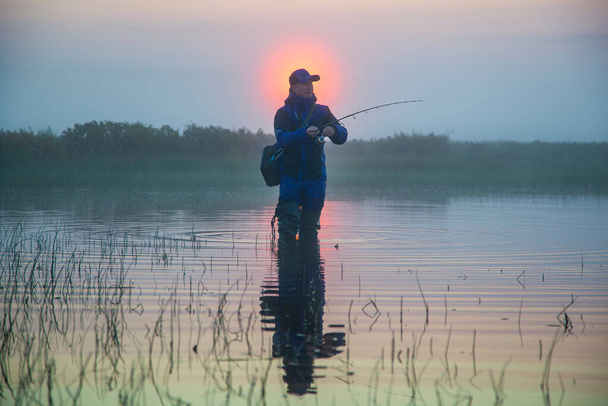 professional fisherman with spinning rod, early morning on the lake, foggy morning over the water, sunrise - Photo, Image
