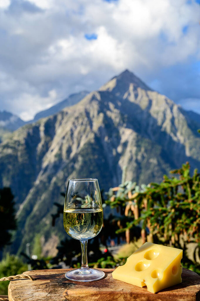 Glass of swiss or savoy dry white wine and yellow emmental cheese with Alpine mountains peaks on background in sunny day - Photo, Image