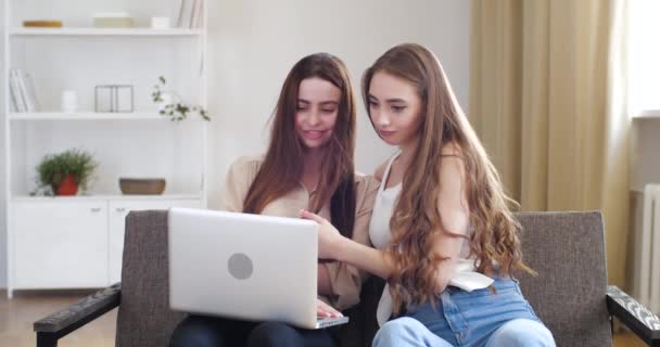 Zwei Teenager-Mädchen sitzen auf der Couch und schauen auf den Laptop-Bildschirm, lesen online, sehen Videos oder Fotos im Netzwerk, umarmen sich und verbringen die Zeit zu Hause zusammen. Schwester tröstet ihren Freund über die Haare streicheln, freundliche Unterstützung - Filmmaterial, Video