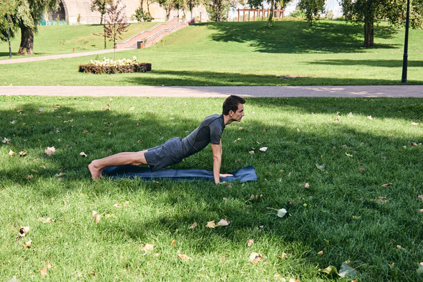 Молодой спортсмен в спортивной одежде занимается йогой в парке. Практика asana на открытом воздухе. Люди тренируются на зеленой траве с ковриком для йоги. Сильный взрослый кавказский мужчина в позе зонтика. - Фото, изображение