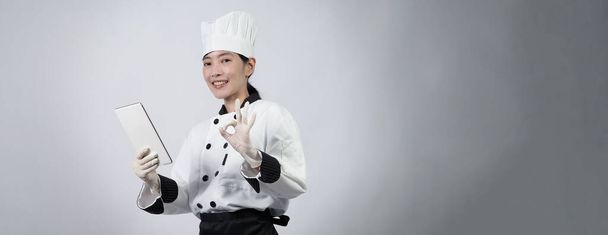 Middle aged of asian woman chef holding smartphone or digital tablet and received order from online shop or merchant application. she smiling in chef uniform and standing in studio with white color background. - Photo, Image