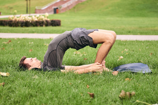 Młody sportowiec w odzieży sportowej uprawiający jogę w parku. Ćwicz asanę na świeżym powietrzu. Ludzie ćwiczący na zielonej trawie z matą do jogi. silny Dojrzałe biały mężczyzna w ramię poza Kandharasana. - Zdjęcie, obraz