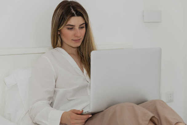 Junge schöne Frau entspannt mit Laptop-Computer auf dem Bett im Schlafzimmer. Online-Chat. Arbeiten von zu Hause aus. Frau checkt soziale Apps. Online Lebensmittel bestellen. Quarantäne bei Coronavirus-Epidemie - Foto, Bild