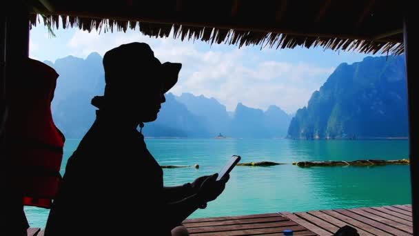 Silueta vista lateral joven turista relajante usando teléfono inteligente en casa balsa de bambú con fondo de montaña de piedra caliza disfrutando de vacaciones de verano en Cheow Lan, Parque Nacional Khao Sok Tailandia - Metraje, vídeo