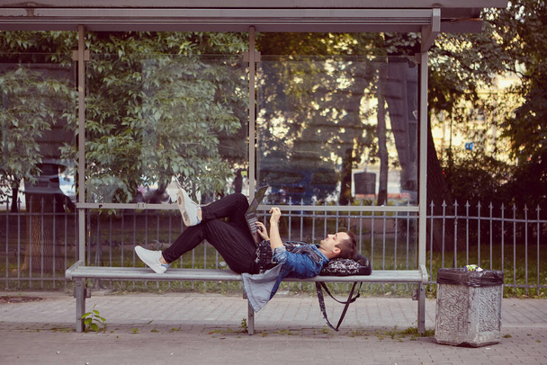 Young man with laptop on his knees is lying on the public transport station while waiting for a bus. - Fotografie, Obrázek