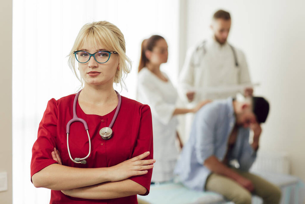 聴診器で赤い制服を着た金髪の女医の肖像画。病院のカメラにポーズをとっている若い医者。背景では二人の医師が患者を診察し - 写真・画像