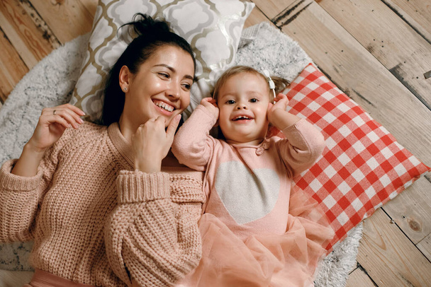 A beautiful brunette woman lies on a warm wooden floor and plays with her daughter. Family having fun together, top view of happy people, motherhood relationship concept - Foto, imagen