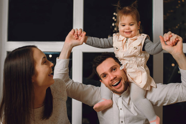 father and mother are having fun with their daughter. The child sits on the shoulders of the father and smiles. The family spends the evening at home by the window showing emotions and a playful mood - Photo, Image