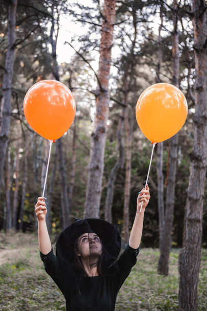 Young cute beautiful woman in dark dress and witchs hat holds orange balloons in her hands. Halloween party costume. Forest, park with autumn trees. - Фото, изображение
