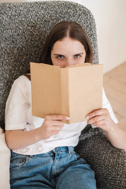 Young woman in jeans and white T-shirt sits in gray armchair and reads paper book. Concept of relaxing holiday, home comfort, hygge, slow-living. - 写真・画像