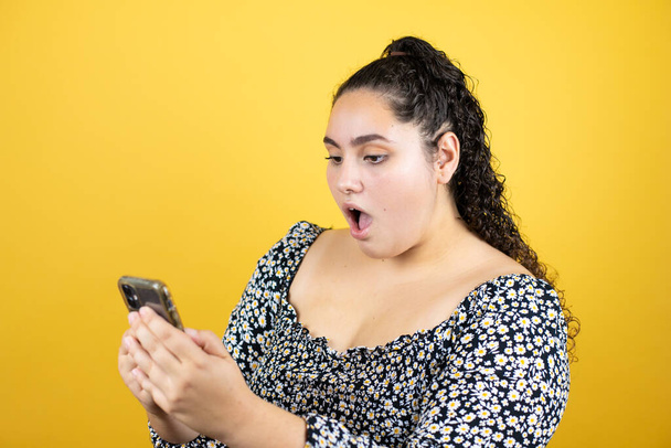 Young beautiful woman with curly hair over isolated yellow background chatting with her phone and surprised - Fotó, kép