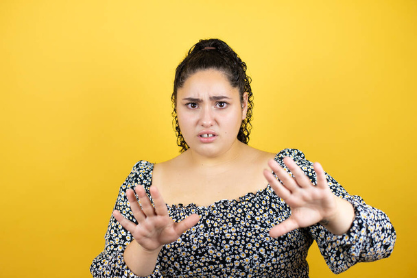 Young beautiful woman with curly hair over isolated yellow background afraid and terrified with fear expression stop gesture with hands, shouting in shock. Panic concept. - Photo, Image