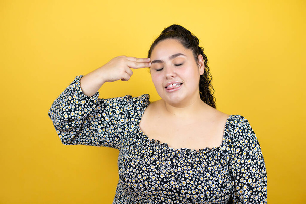Young beautiful woman with curly hair over isolated yellow background hooting and killing oneself pointing hand and fingers to head like gun, suicide gesture. - Photo, Image