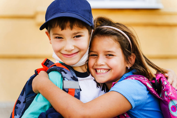 Small boy and girl caucasian brother and sister or little friends hug with backpacks on their shoulders going back to school - Little first graders in summer day close up portrait smiling - Photo, Image