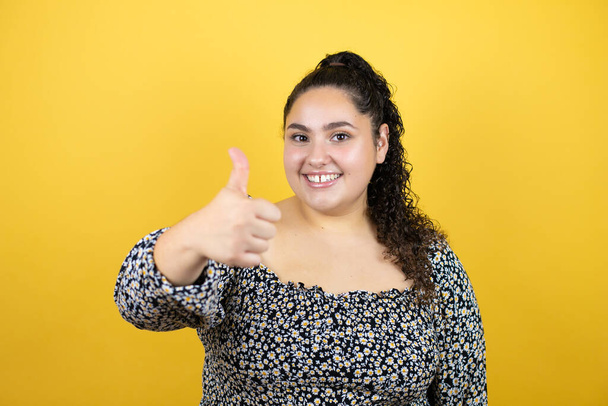 Young beautiful woman with curly hair over isolated yellow background smiling and doing the ok signal with her thumb - Photo, Image
