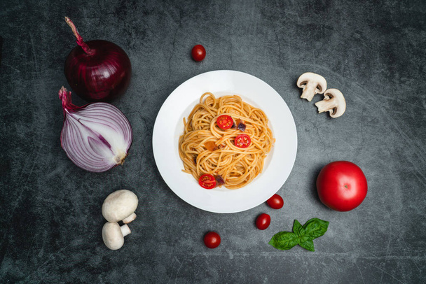 Heerlijke tomaat en paddestoel spaghetti op een wit plateEen bord spaghetti en ingrediënten op een donkergrijze achtergrond - Foto, afbeelding