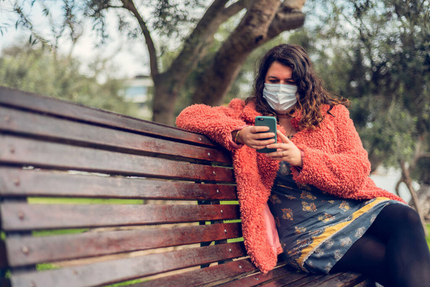 Tıbbi maskeli güçlü bir kadın akıllı telefonuna dışarıdaki parkta bir bankta oturuyor. Yeni normal konsept. Boşluğu kopyala - Fotoğraf, Görsel