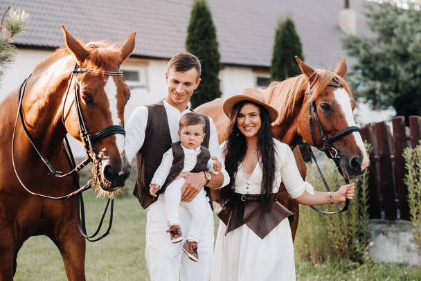 A family in white clothes with their son stand near two beautiful horses in nature. A stylish couple with a child are photographed with horses. - Foto, Imagem