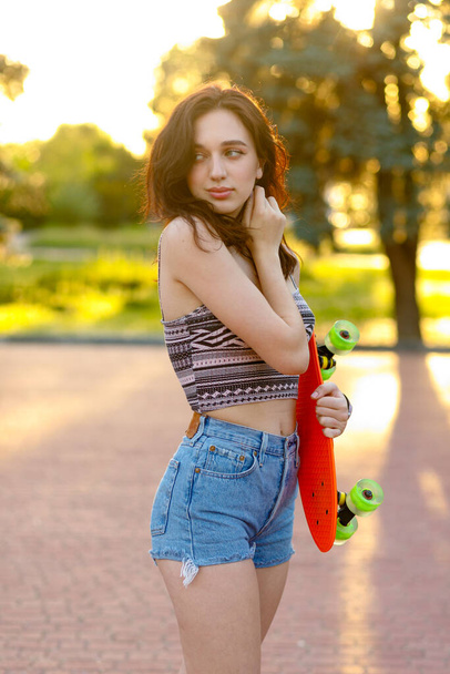 Beautiful active brunette girl with long hair wearing top, blue denim shorts and stylish pink sneakers posing on sunset background. holding a orange skate with green wheels. - Фото, зображення