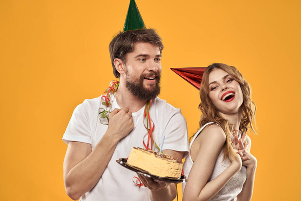Happy man and woman in a cap celebrating a birthday on a yellow background with a cake in their hands - Φωτογραφία, εικόνα