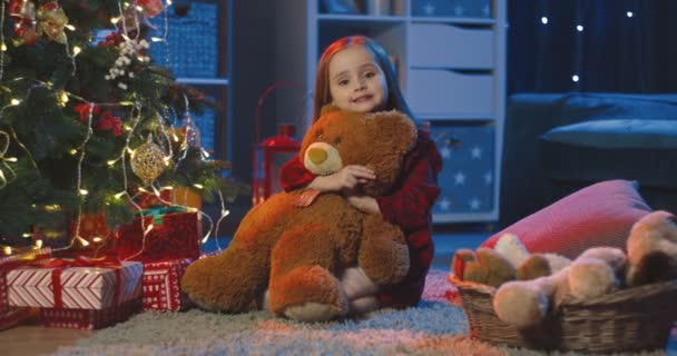 Muotokuva kaunis ja pieni tyttö istuu lattialla, halaus nallekarhu ja hymyillen kameran pimeässä huoneessa x-mas puu jouluna. - Materiaali, video