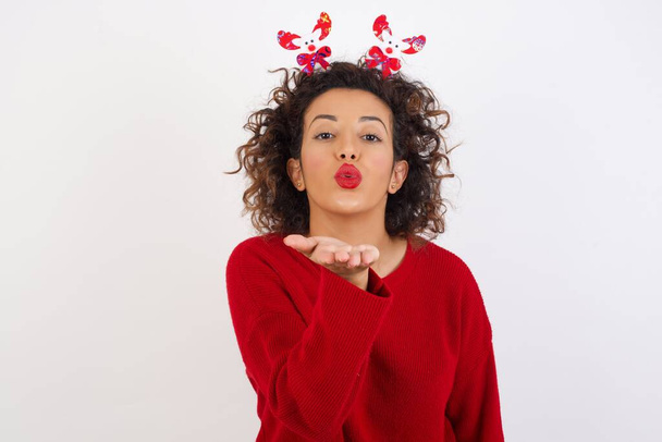 woman with curly hair wearing deer horns headband, looking at the camera blowing a kiss with hand, love kissing  - Photo, Image
