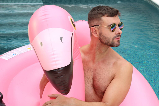 Yüzme havuzunda pembe şişme flamingo ile kameraya bakan yakışıklı genç adamın yakın plan portresi. - Fotoğraf, Görsel