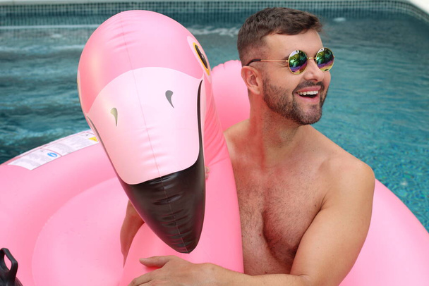 Yüzme havuzunda pembe şişme flamingo ile kameraya bakan yakışıklı genç adamın yakın plan portresi. - Fotoğraf, Görsel