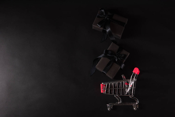 Black Friday sale shopping concept, Top view of gift box wrapped black paper and black bow ribbon present dropping to the shopping cart, studio shot on black background - Fotó, kép