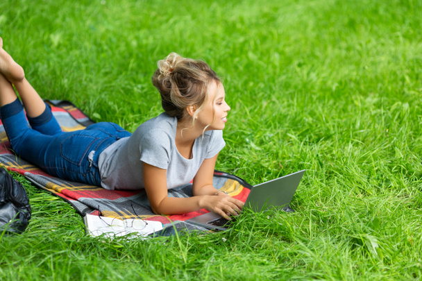 Young woman - watching educational webinar on laptop, lying on green grass. Online meeting with friends. - Foto, afbeelding