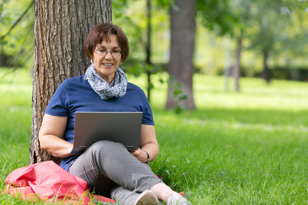 Sorridente donna anziana che lavora a distanza su un portatile nel parco cittadino. Concetto di lavoro distante - Foto, immagini
