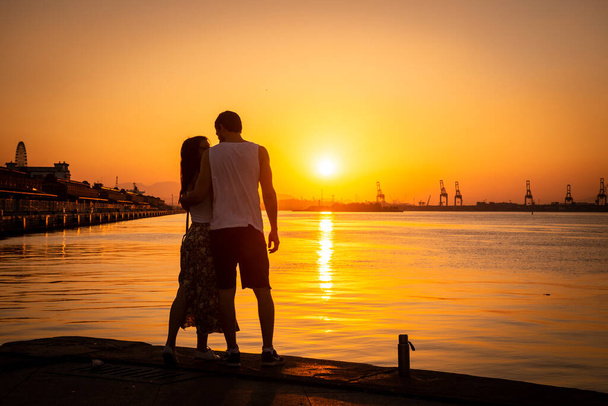 Beautiful view to young adult couple watching the sunset by the ocean city shore in Guanabara Bay, Rio de Janeiro, Brazil - Fotoğraf, Görsel