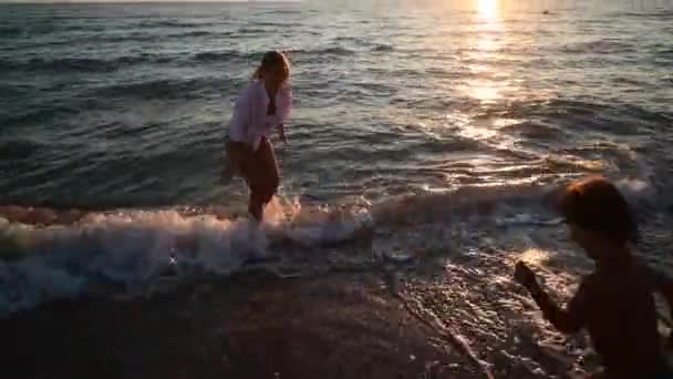 Mujer joven y su hijo vienen al agua en la puesta de sol en la playa de arena, vacaciones en la isla - Metraje, vídeo