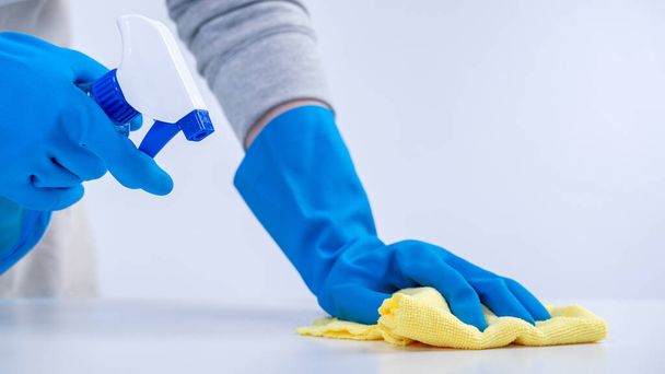 Young woman housekeeper in apron is cleaning, wiping down table surface with blue gloves, wet yellow rag, spraying bottle cleaner, closeup design concept. - Photo, Image