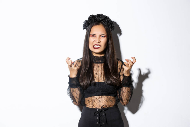 Image of angry wicked witch cursing and swearing, clenching fists and grimacing with hatred, standing over white background, celebrating halloween - Fotó, kép