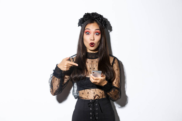Портрет изумленной азиатской девушки, указывающей пальцем на смартфон, показывающей что-то о Хэллоуине в интернете, стоящей на белом фоне в готическом кружевном платье - Фото, изображение