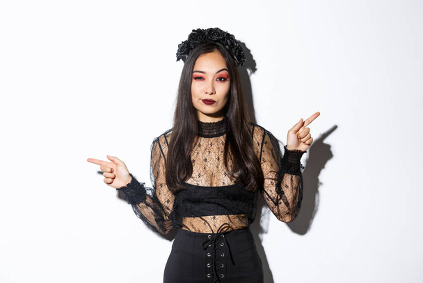 Sassy young evil witch with gothic makeup and wreath, looking arrogant while pointing fingers sideways, showing two halloween themed banners, standing over white background - Photo, Image