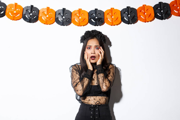 Bild einer Frau im Hexenkostüm, die erschrocken schaut, Schrecken oder Angst ausdrückt, während sie über weißem Hintergrund mit Kürbisfahnen-Dekoration steht und Halloween feiert - Foto, Bild